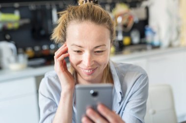 Evde mutfaktaki neşeli, neşeli genç kadın cep telefonundaki sosyal medya uygulamalarını sevdikleriyle sohbet etmek ve casusluk yapmak için kullanıyor. Evde kal, sosyal mesafeli yaşam tarzı.