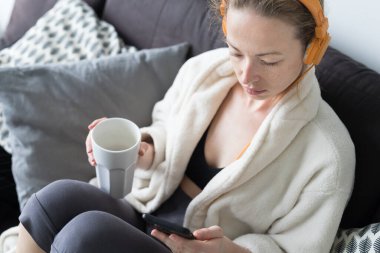 Evde kal. Sosyal uzaklık. Kanepede uzanan, beyaz bardaktan çay içen, rahatlatıcı müzik dinleyen, mobil ağlar üzerinden aile ve arkadaşlarıyla iletişim kuran bir kadın..