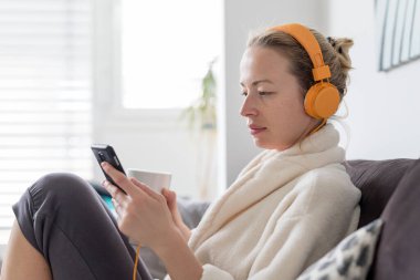 Evde kal. Sosyal uzaklık. Kanepede uzanan, beyaz bardaktan çay içen, rahatlatıcı müzik dinleyen, mobil ağlar üzerinden aile ve arkadaşlarıyla iletişim kuran bir kadın..