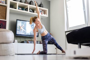 Evde spor yapan çekici bir kadın oturma odasında televizyon karşısında pilates egzersizi yapıyor. Sosyal uzaklık. Sağlıklı kalın ve Corona virüsü salgınında evde kalın.