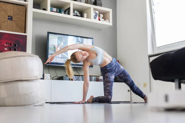 Atrakcyjna sportowa kobieta ćwicząca w domu, ćwicząca pilates przed telewizorem w swoim salonie. Oddalenie się od społeczeństwa. Bądź zdrowy i pozostań w domu podczas pandemii wirusa korony — Zdjęcie stockowe