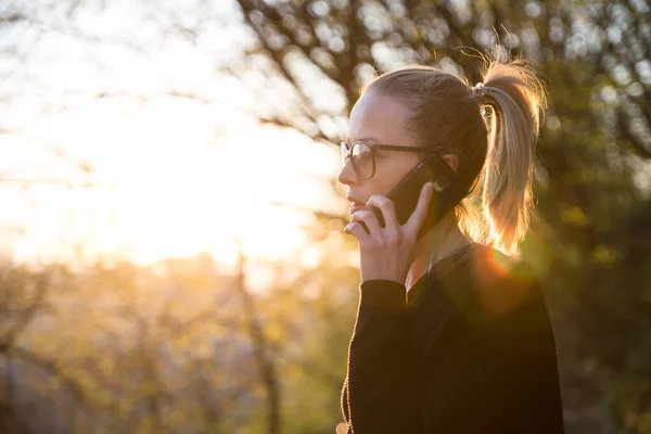 Backlit rear view of young woman talking on cell phone outdoors in park at sunset. Girl holding mobile phone, using digital device, looking at setting sun — Stock Photo, Image