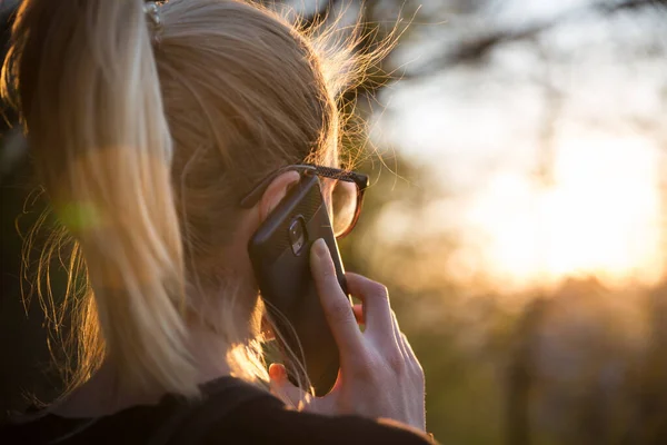 Bakgrundsbelyst bakre vy av ung kvinna talar på mobiltelefon utomhus i parken vid solnedgången. Flicka hålla mobiltelefon, med hjälp av digital enhet, titta på nedgående sol — Stockfoto