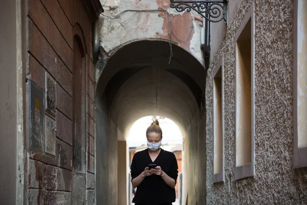 Coronavirus pandémico COVID-19. Mujer caucásica casual en la calle de la ciudad medieval usando teléfono móvil, usando mascarilla protectora contra la propagación del coronavirus y la transmisión de enfermedades — Foto de Stock