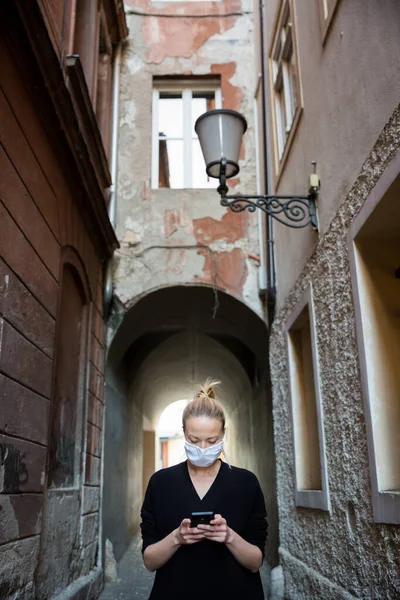 Coronavirus pandemico COVID-19. Casual donna caucasica in strada città medievale utilizzando il telefono cellulare, indossando maschera protettiva contro la diffusione del coronavirus e la trasmissione della malattia — Foto Stock