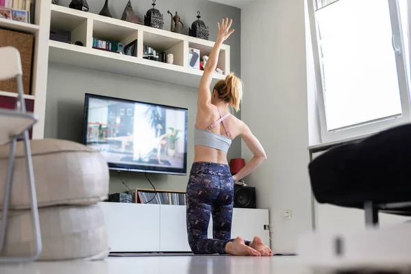 Atrakcyjna sportowa kobieta ćwicząca w domu, ćwicząca pilates przed telewizorem w swoim salonie. Oddalenie się od społeczeństwa. Bądź zdrowy i pozostań w domu podczas pandemii wirusa korony — Zdjęcie stockowe