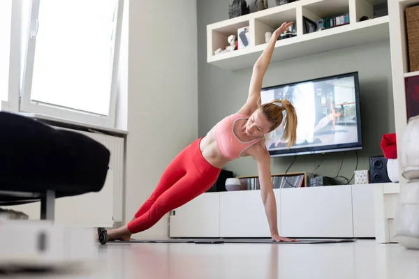 Atrakcyjna sportowa kobieta ćwicząca w domu, ćwicząca pilates przed telewizorem w swoim salonie. Oddalenie się od społeczeństwa. Bądź zdrowy i pozostań w domu podczas pandemii wirusa korony — Zdjęcie stockowe