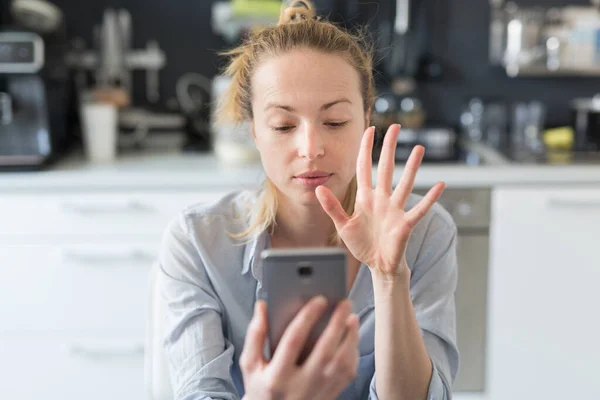 Ung nöjd kvinna inomhus i köket med hjälp av sociala medier appar på mobilen för att chatta och sticka i samband med sina nära och kära. Stanna hemma, social distanserande livsstil. — Stockfoto