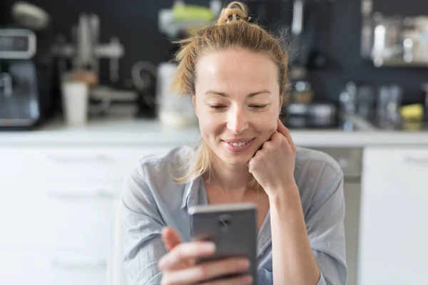 Jovem sorrindo alegre mulher satisfeita dentro de casa cozinha usando aplicativos de mídia social no telefone móvel para conversar e ficar conectado com seus entes queridos. Fique em casa, estilo de vida de distanciamento social . — Fotografia de Stock