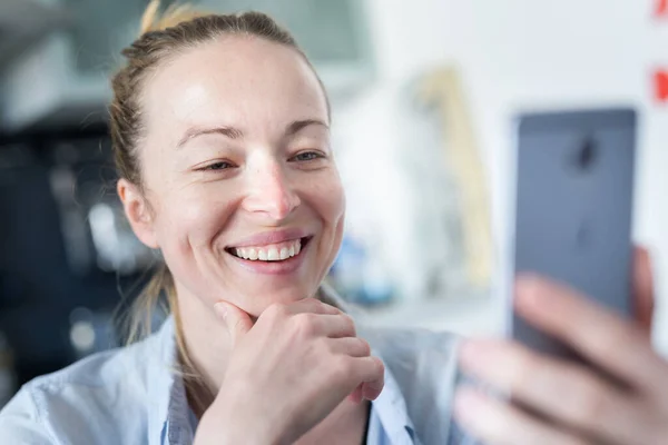 Jovem sorrindo alegre mulher satisfeita dentro de casa cozinha usando aplicativos de mídia social no telefone móvel para conversar e ficar conectado com seus entes queridos. Fique em casa, estilo de vida de distanciamento social . — Fotografia de Stock