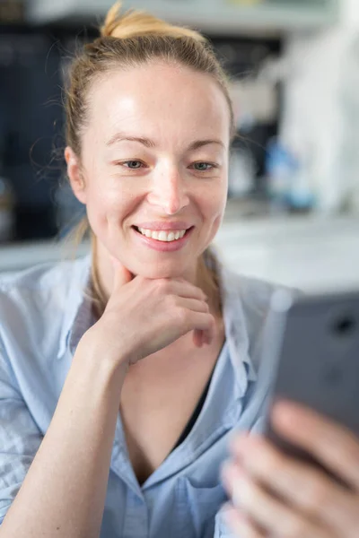 Unga leende glad nöjd kvinna inomhus i köket med hjälp av sociala medier apps på mobiltelefon för att chatta och sticka i samband med sina nära och kära. Stanna hemma, social distanserande livsstil. — Stockfoto