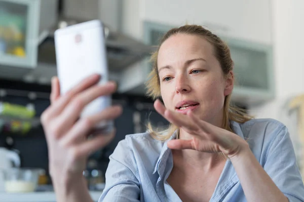 Νεαρή ικανοποιημένη γυναίκα σε εσωτερικούς χώρους στο σπίτι κουζίνα χρησιμοποιώντας εφαρμογές κοινωνικής δικτύωσης στο κινητό τηλέφωνο για συνομιλία και stying συνδέεται με τους αγαπημένους της. Μείνετε στο σπίτι, κοινωνική ζωή αποστασιοποίηση. — Φωτογραφία Αρχείου