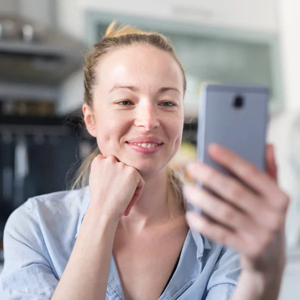 Unga leende glad nöjd kvinna inomhus i köket med hjälp av sociala medier apps på mobiltelefon för att chatta och sticka i samband med sina nära och kära. Stanna hemma, social distanserande livsstil. — Stockfoto