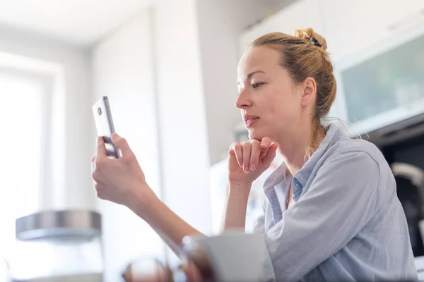 Jeune femme heureuse souriante à l'intérieur de la cuisine à la maison en utilisant les médias sociaux sur le téléphone mobile pour discuter et stying connecté avec ses proches. Rester à la maison, mode de vie social distanciant. — Photo