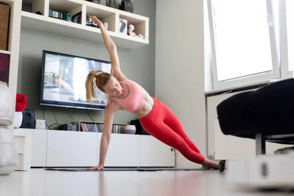 Atrakcyjna sportowa kobieta ćwicząca w domu, ćwicząca pilates przed telewizorem w swoim salonie. Oddalenie się od społeczeństwa. Bądź zdrowy i pozostań w domu podczas pandemii wirusa korony — Zdjęcie stockowe