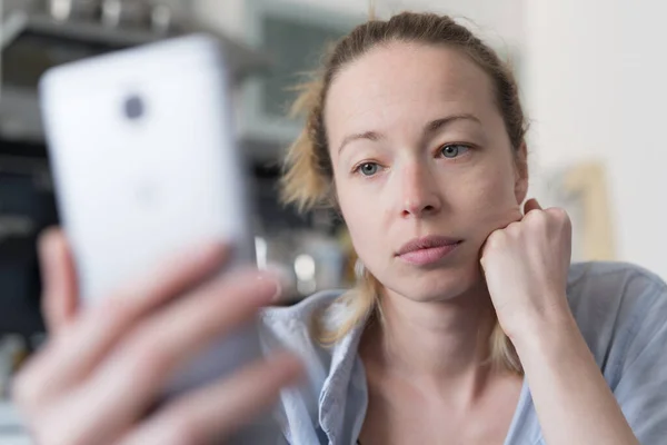 Evde mutfaktaki endişeli kadın telefonda sosyal medya uygulamalarını kullanarak görüntülü konuşma ve sevdikleriyle casusluk yapıyor. Evde kal, sosyal mesafeli yaşam tarzı — Stok fotoğraf