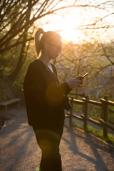 Gün batımında parkta cep telefonuyla konuşan genç bir kadının arka plan görüntüsü. Cep telefonu tutan, dijital cihaz kullanan, batan güneşe bakan kız — Stok fotoğraf