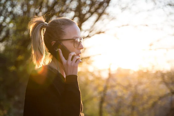 Backlit rear view of young woman talking on cell phone outdoors in park at sunset. Girl holding mobile phone, using digital device, looking at setting sun — Stock Photo, Image