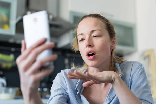 Ung nöjd kvinna inomhus i köket med hjälp av sociala medier appar på mobilen för att chatta och sticka i samband med sina nära och kära. Stanna hemma, social distanserande livsstil. — Stockfoto