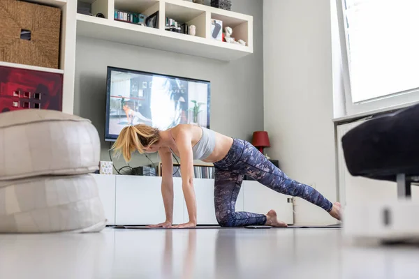 Atrakcyjna sportowa kobieta ćwicząca w domu, ćwicząca pilates przed telewizorem w swoim salonie. Oddalenie się od społeczeństwa. Bądź zdrowy i pozostań w domu podczas pandemii wirusa korony — Zdjęcie stockowe