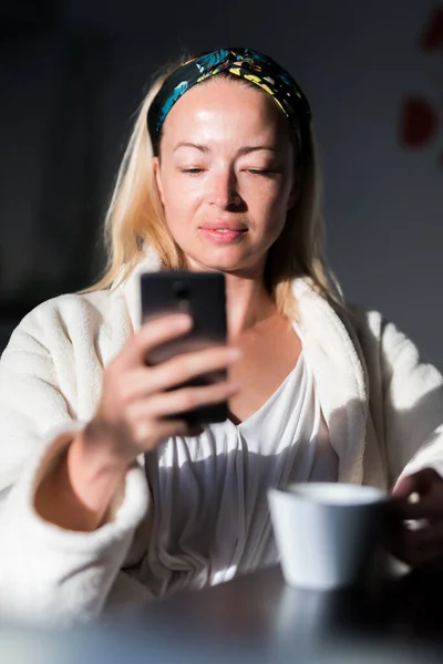 Vacker kaukasiska kvinna hemma, känna sig bekväm klädd i vit badrock, ta lite tid för sig själv, dricka morgonkaffe och läsa nyheter på mobiltelefon enhet på morgonen — Stockfoto