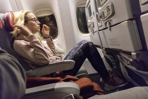 Sporty casual young blonde caucasian lady watching movie while traveling by airplane by the window. Commercial transportation by planes — Stock Photo, Image