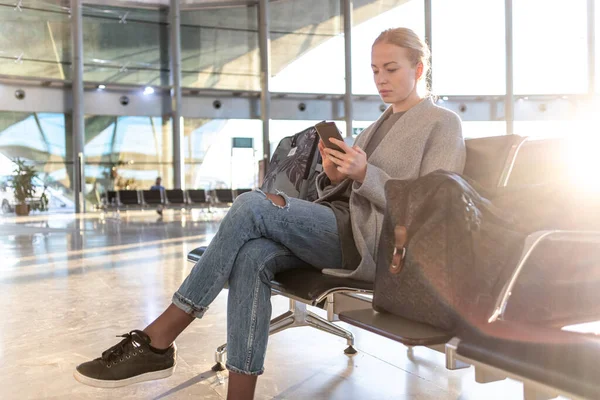 Jovem loira casual usando seu celular enquanto espera para embarcar em um avião nos portões de partida do aeroporto. Terminal do aeroporto vazio devido à pandemia do vírus da corona — Fotografia de Stock