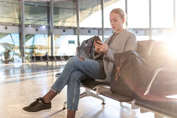 Zwykła blondynka używająca komórki, czekając na wsiadanie do samolotu przy wyjściu z lotniska. Pusty terminal lotniska z powodu pandemii wirusa korony — Zdjęcie stockowe