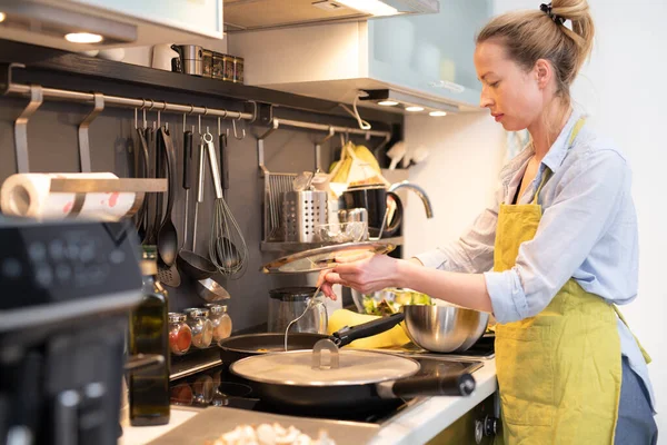 Stay at home housewife woman cooking in kitchen, stir frying dish in a saucepan, preparing food for family dinner.