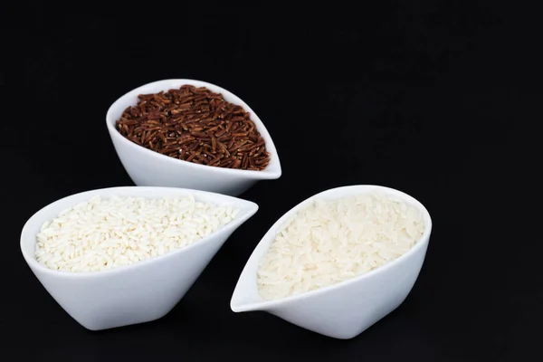 3 tipos de arroz tailandés crudo Arroz de jazmín tailandés (Hom Mali), Arroz glutinoso / pegajoso (Khao Neow), Arroz integral (Khao Dum) en tazas blancas sobre fondo negro con espacio para copiar — Foto de Stock