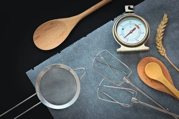 Pékség előkészítő eszközök fakanállal, vanília, hőmérő, sütni papír, fekete háttér — Stock Fotó