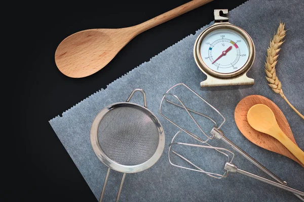 Хлебопекарные инструменты деревянная ложка, ваниль, термометр, бумага для выпечки на черном фоне — стоковое фото