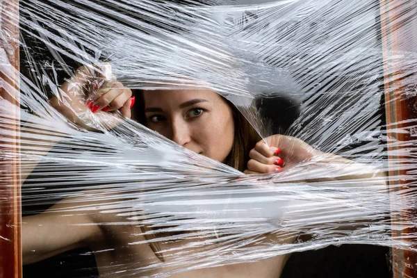 mysterious portrait girl hidden white behind breaking stretch cellophane and looking through the gap plastic. Pretty brunette wrapped in cellophane.