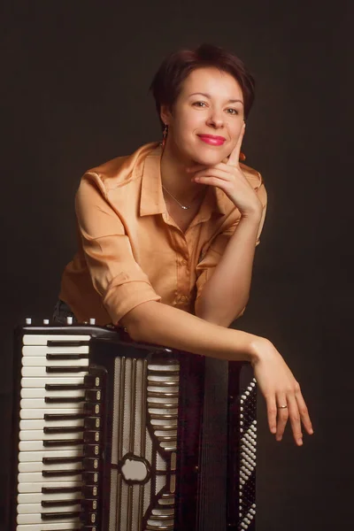 A brunette in a gold shirt, with short haircut, on a dark brownbackground of the Studio. With accordion musician