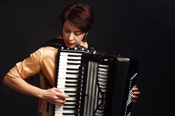 A brunette in a gold shirt, with a short haircut, on a dark background of the Studio. play the accordion