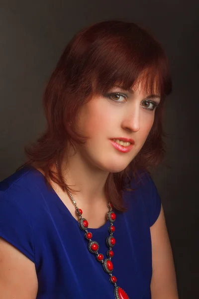Portrait of a full grown woman, brunette, in the Studio on a gray background. In a blue dress and red jewelry