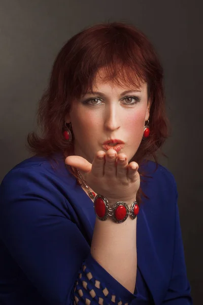 Portrait of a full grown woman, brunette, in the Studio on a gray background. In a blue dress and red jewelry. sends an air kiss