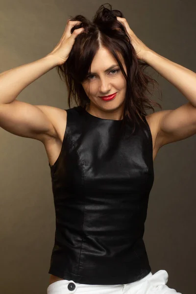 a brunette in a black leather sleeveless blouse and white trousers poses on a gray background. warm toning