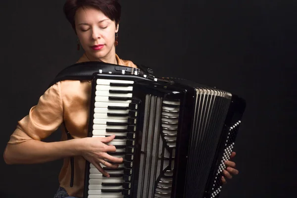 A brunette in a gold shirt, with a short haircut, on a dark background of the Studio. play the accordion