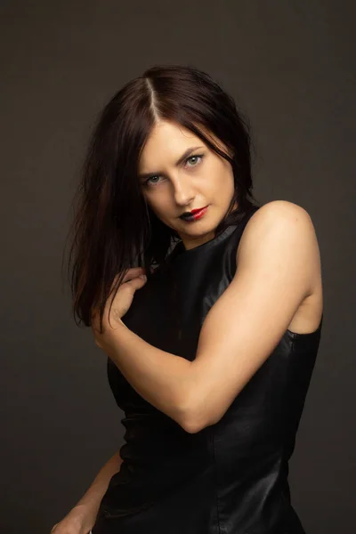 a brunette in a black leather sleeveless blouse poses on a gray background. warm toning.lips painted black and red