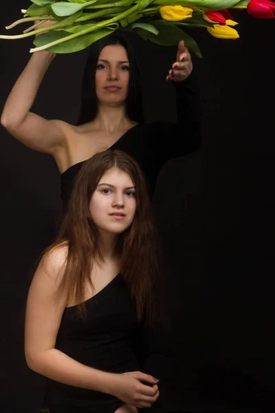 Portrait of girl and woman, mother and daughter, in a black one-shoulder dress open, naked , with red and yellow tulips, in Studio on black background
