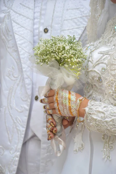 Indonesian bride's hand holding a flower bouquet, white kebaya and white henna on the hand