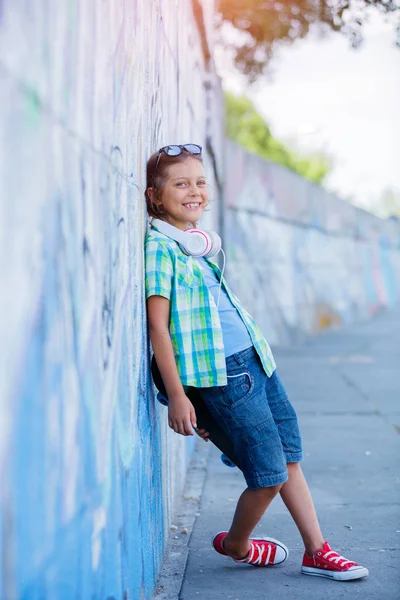 Roztomilý chlapec s skateboard venku, stojící na ulici s různými barevnými graffiti na zdi — Stock fotografie