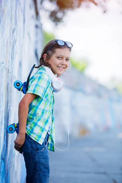 Schattige jongen met skateboard buitenshuis, staande op de straat met verschillende kleurrijke graffiti op de muren — Stockfoto