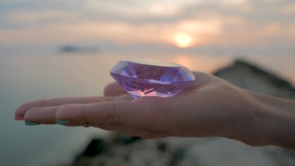 Purple diamond crystal in a female hand, close up, on sunset background — Stock Video