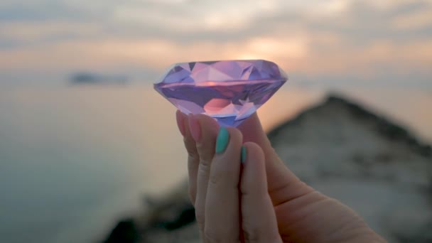 Purple diamond crystal in a female hand, close up, on sunset background — Stock Video