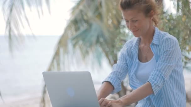 Young woman sitting at the table with a laptop with sea view behind — Stock Video