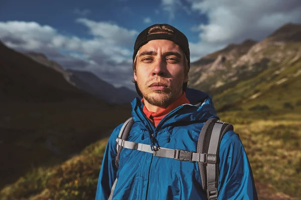 Mladý turista v modré bundě s batohem stojící a dívající se do kamery v horách — Stock fotografie