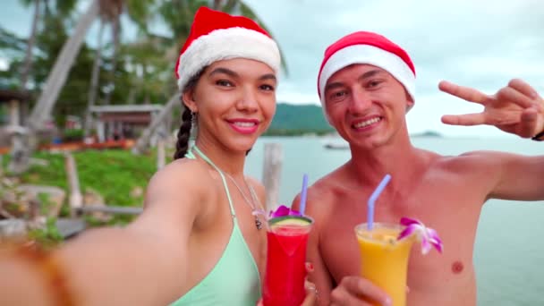 Счастливая пара улыбается перед камерой, снимая автопортрет в шляпах Санты на пляже — стоковое видео