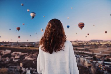 Dağın tepesinde beyaz kazak giyen turist bir kız Cappadocia 'da balon manzarasının tadını çıkarıyor. Türkiye 'de İyi Yolculuklar konsepti